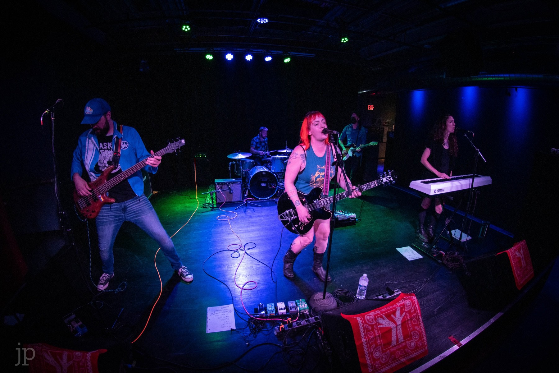 The Cancellations, a rock band, plays a song on a large stage. There is a bassist, a guitarist and vocalist with orange hair, and a keyboard player. The stage is lit in shades of blue and purple.