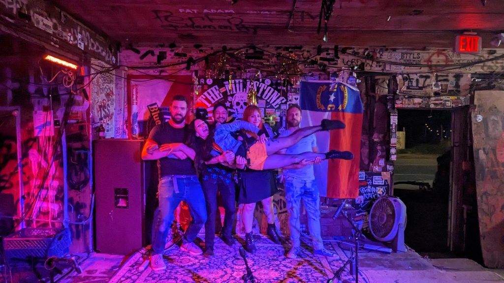 The Cancellations stand on stage at The Milestone Club in front of the venue's logo. The rest of the band is holding Kerri up in the air.