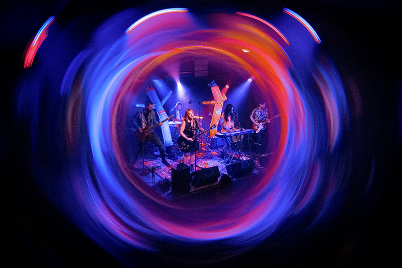 The Cancellations play live on stage at a concert. The center of the photo is framed by slow exposure lights of blue, orange and pink.