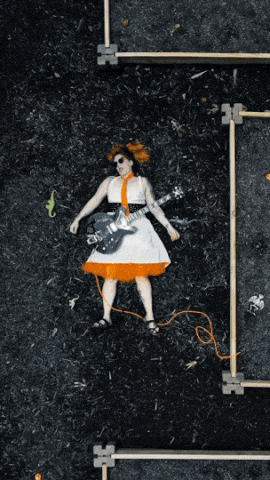 A looping video of Ellie laying in the dirt playing guitar and pretending to die on repeat, shot from above. She is wearing a white dress with an orange tie and crinoline.