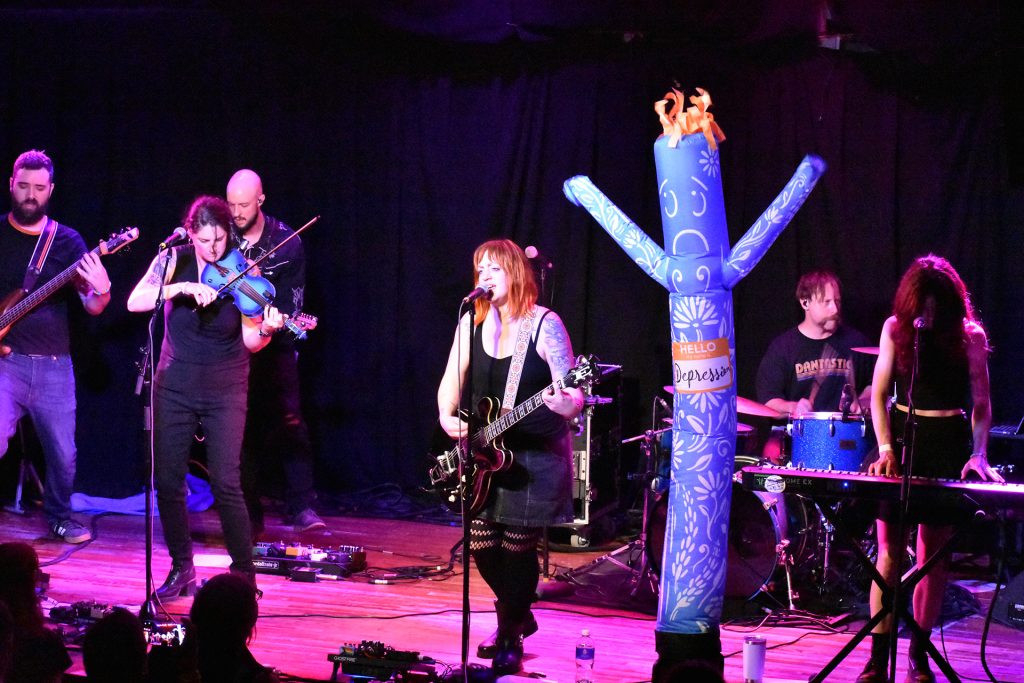 The Cancellations play live on stage at the High Noon in Madison. The stage is lit in magenta lights. Depression, the inflatable flailing tube mascot, dances between Ellie and Gary.